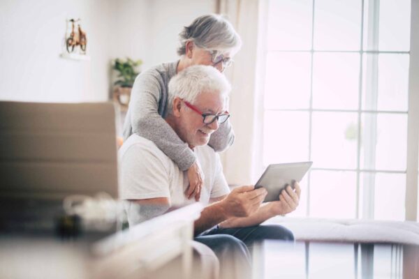 Couple,Of,Seniors,Smiling,And,Looking,At,The,Same,Tablet