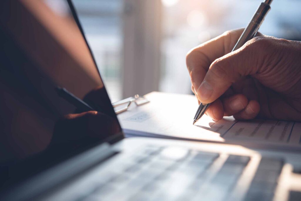 Person writing notes by a laptop