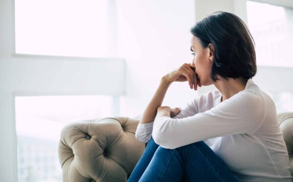 Close-up photo of an adult woman who turned away from the camera, looking through the window with sadness.