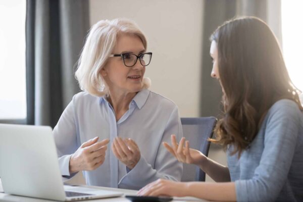 Mature,And,Young,Women,Colleagues,Sitting,At,Desk,Talking,About