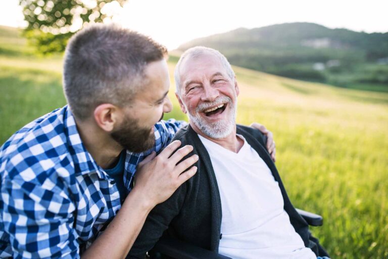 An adult son with senior father in wheelchair on a walk in nature at sunset, laughing.
